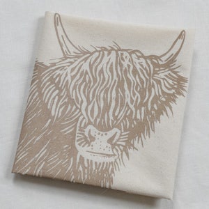 May include: Folded beige tea towel featuring a detailed illustration of a Highland cow's face in shades of brown. The cow's long fur and horns are prominently displayed, creating a rustic and charming design. The towel is made of a soft, natural fabric.