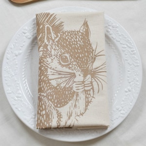May include: A white linen napkin with a brown squirrel illustration printed on it. The squirrel is holding a nut in its mouth. The napkin is folded and placed on a white plate.