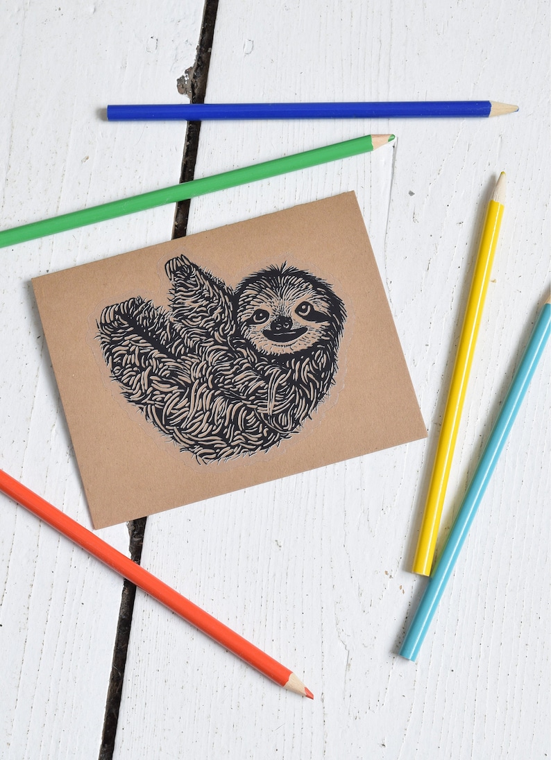 May include: A brown card with a black and white illustration of a sloth on it. The card is surrounded by colored pencils on a white wooden surface.