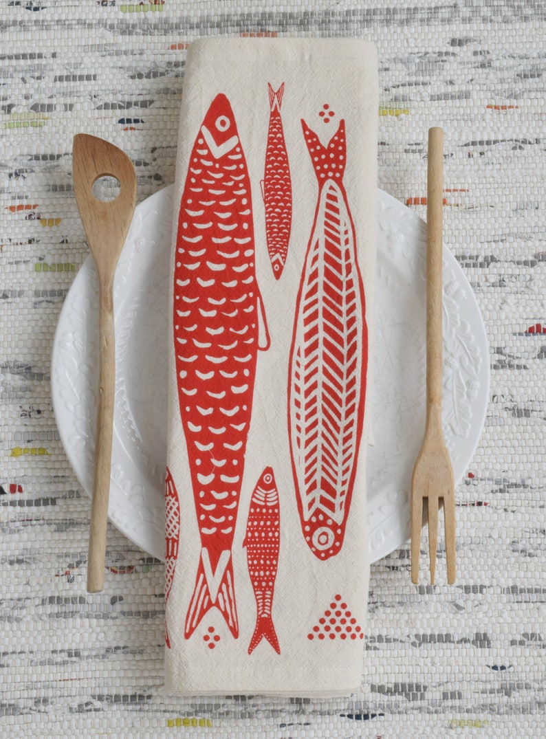 May include: A white linen napkin with a red fish design. The napkin is folded and placed on a white plate with a wooden spoon and fork.