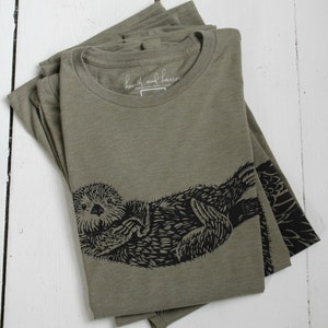 May include: A folded green t-shirt with a black and white illustration of a sea otter. The shirt has a label that reads "heath and harrow".