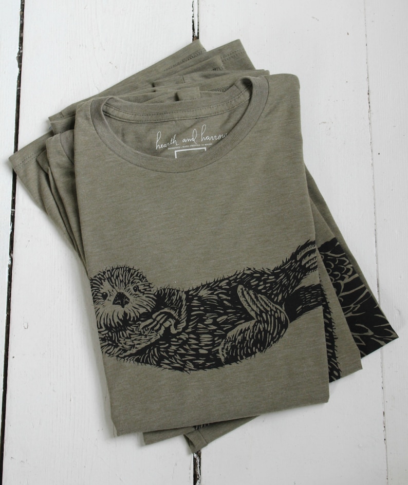 May include: A folded green t-shirt with a black and white illustration of a sea otter. The shirt has a label that reads "heath and harrow".