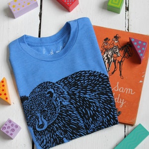 May include: A blue t-shirt with a black bear graphic print. The shirt has a crew neck and short sleeves. The text "Little and Large" is visible on the shirt.