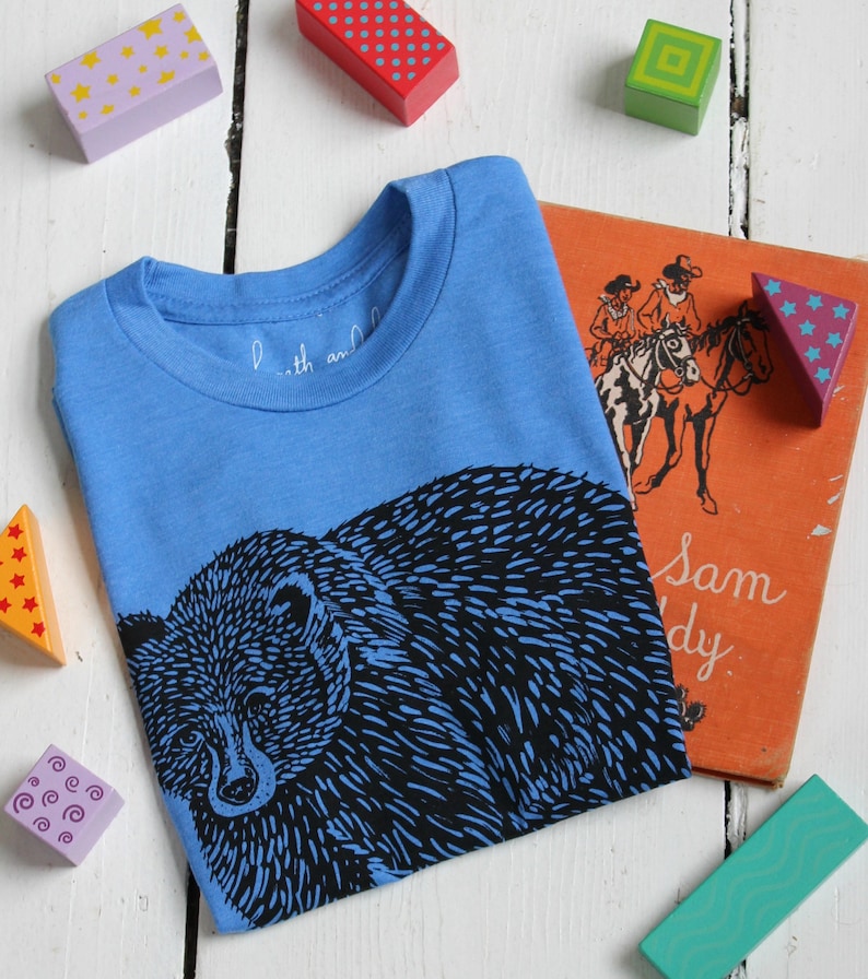 May include: A blue t-shirt with a black bear graphic print. The shirt has a crew neck and short sleeves. The text "Little and Large" is visible on the shirt.