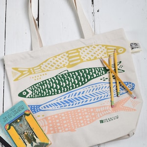 May include: Cream-colored tote bag with four colorful fish illustrations in yellow, green, blue, and pink. The bag has cream-colored handles and the text "HEARTH & HARROW". Pencils and a book are next to the bag.
