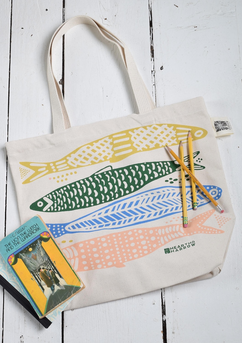 May include: Cream-colored tote bag with four colorful fish illustrations in yellow, green, blue, and pink. The bag has cream-colored handles and the text "HEARTH & HARROW". Pencils and a book are next to the bag.