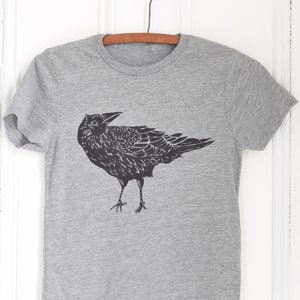 Pode incluir: Uma camiseta cinza com uma ilustra&ccedil;&atilde;o preta de um corvo. O corvo &eacute; retratado de forma detalhada e estilizada, com suas asas e penas cuidadosamente representadas. A camiseta est&aacute; pendurada em um cabide de madeira sobre um fundo branco.