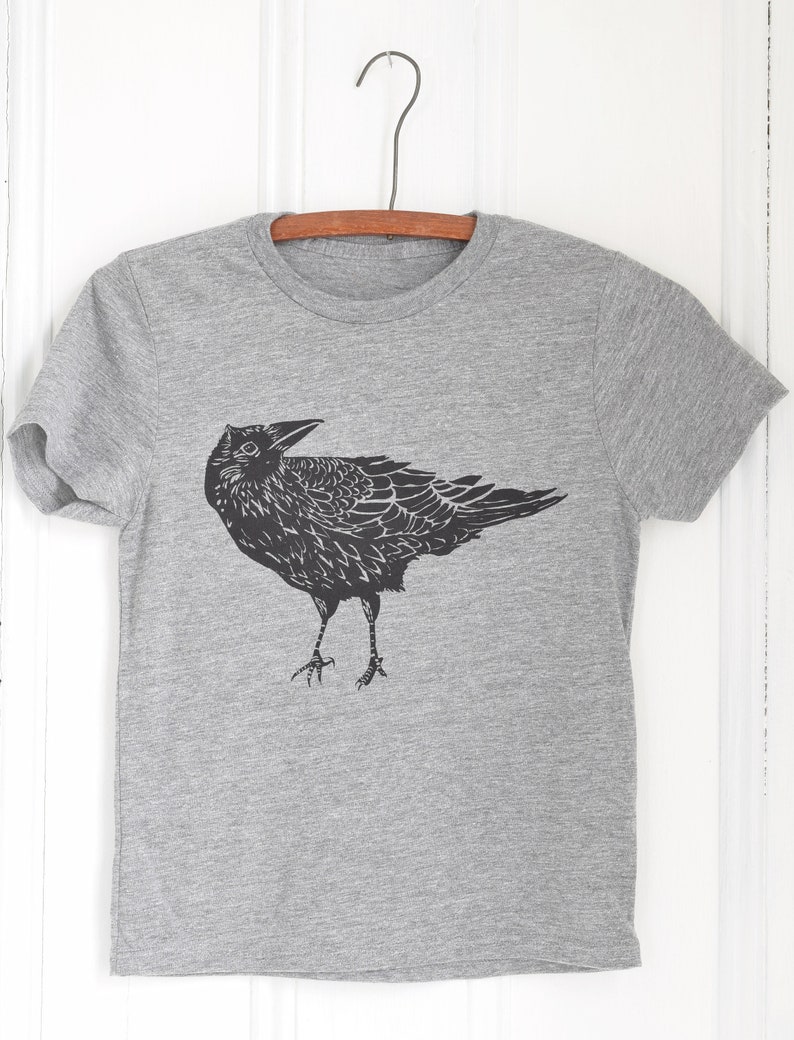Pode incluir: Uma camiseta cinza com uma ilustra&ccedil;&atilde;o preta de um corvo. O corvo &eacute; retratado de forma detalhada e estilizada, com suas asas e penas cuidadosamente representadas. A camiseta est&aacute; pendurada em um cabide de madeira sobre um fundo branco.