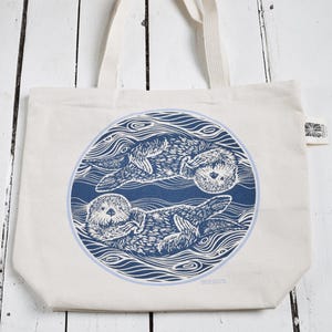 May include: Cream-colored tote bag featuring a blue circular design with two otters floating in water. The otters are depicted in a detailed, linocut style. The bag has two cream-colored handles.