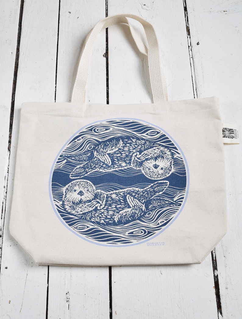May include: Cream-colored tote bag featuring a blue circular design with two otters floating in water. The otters are depicted in a detailed, linocut style. The bag has two cream-colored handles.