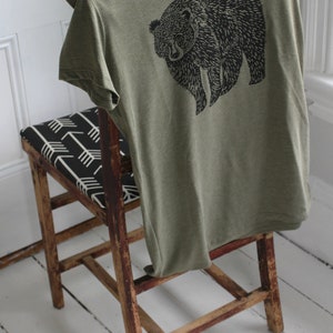K&ouml;nnte beinhalten: Ein gr&uuml;nes T-Shirt mit einem schwarzen B&auml;ren-Motiv auf der Vorderseite. Das T-Shirt ist gefaltet und &uuml;ber einen Holzstuhl mit einem schwarz-wei&szlig;en gemusterten Sitz gelegt.