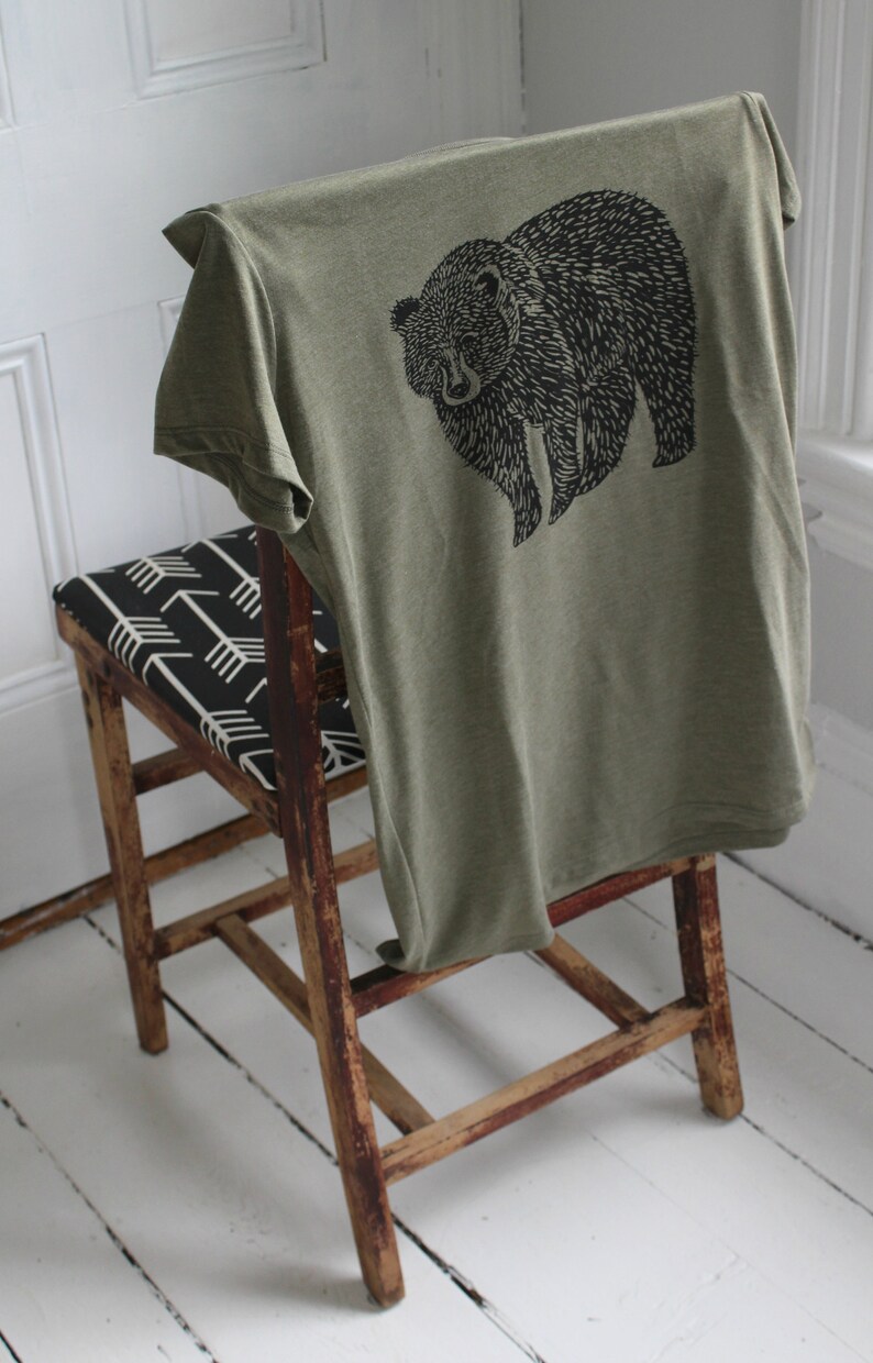 K&ouml;nnte beinhalten: Ein gr&uuml;nes T-Shirt mit einem schwarzen B&auml;ren-Motiv auf der Vorderseite. Das T-Shirt ist gefaltet und &uuml;ber einen Holzstuhl mit einem schwarz-wei&szlig;en gemusterten Sitz gelegt.
