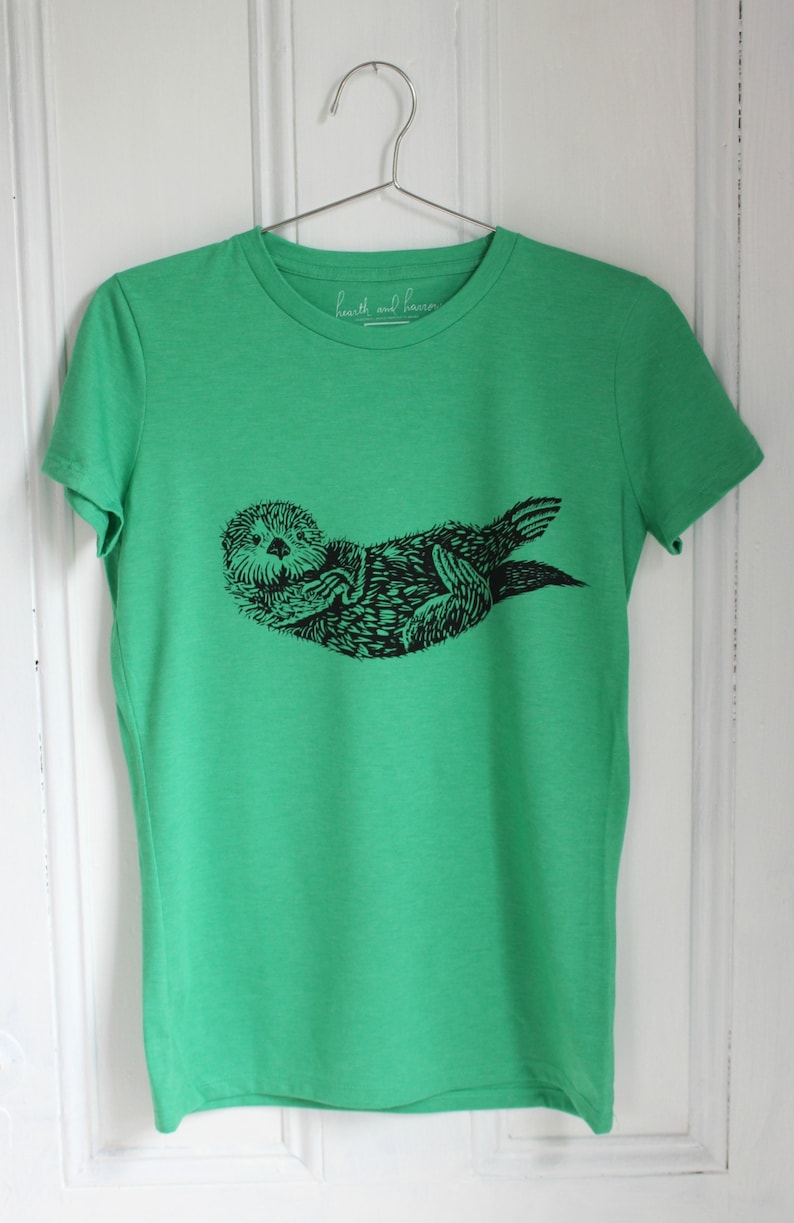 K&ouml;nnte beinhalten: Gr&uuml;nes T-Shirt mit einer schwarzen Grafik einer Seeotter, die auf dem R&uuml;cken liegt. Das Shirt hat einen Rundhalsausschnitt und kurze &Auml;rmel. Der Text "hearth and harbor" ist auf dem Ausschnitt aufgedruckt.