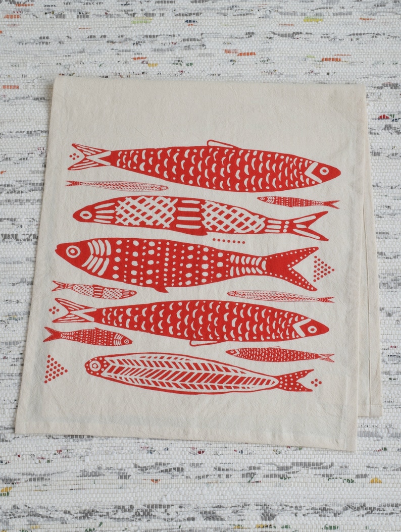 May include: A white tea towel with a red print of various sized fish. The fish are stylized and have different patterns, including dots, lines, and scales.