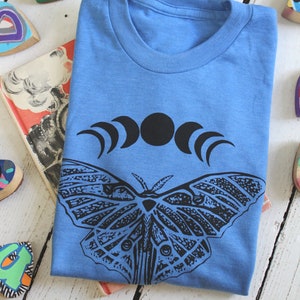 May include: A light blue t-shirt with a black and white graphic of a moth and a crescent moon design.