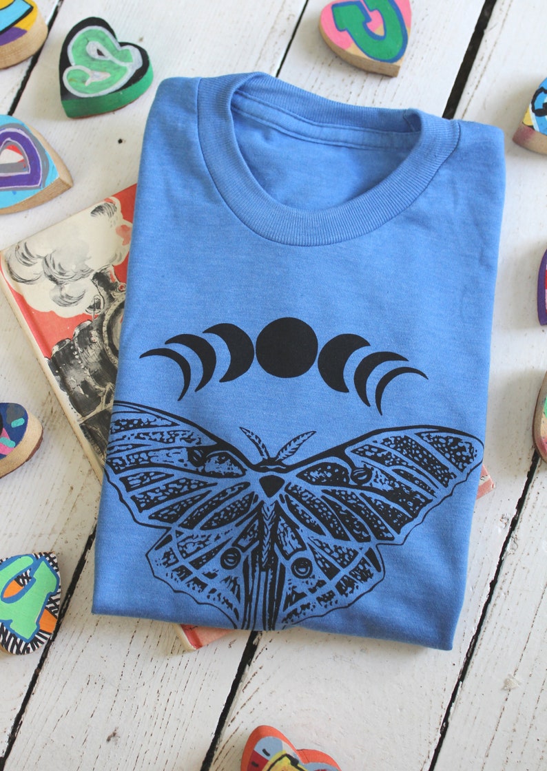 May include: A light blue t-shirt with a black and white graphic of a moth and a crescent moon design.