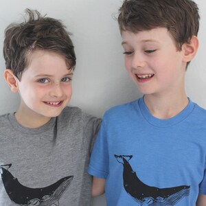 May include: Two boys wearing t-shirts with a whale graphic. The boy on the left is wearing a gray t-shirt with a black whale graphic. The boy on the right is wearing a blue t-shirt with a black whale graphic.