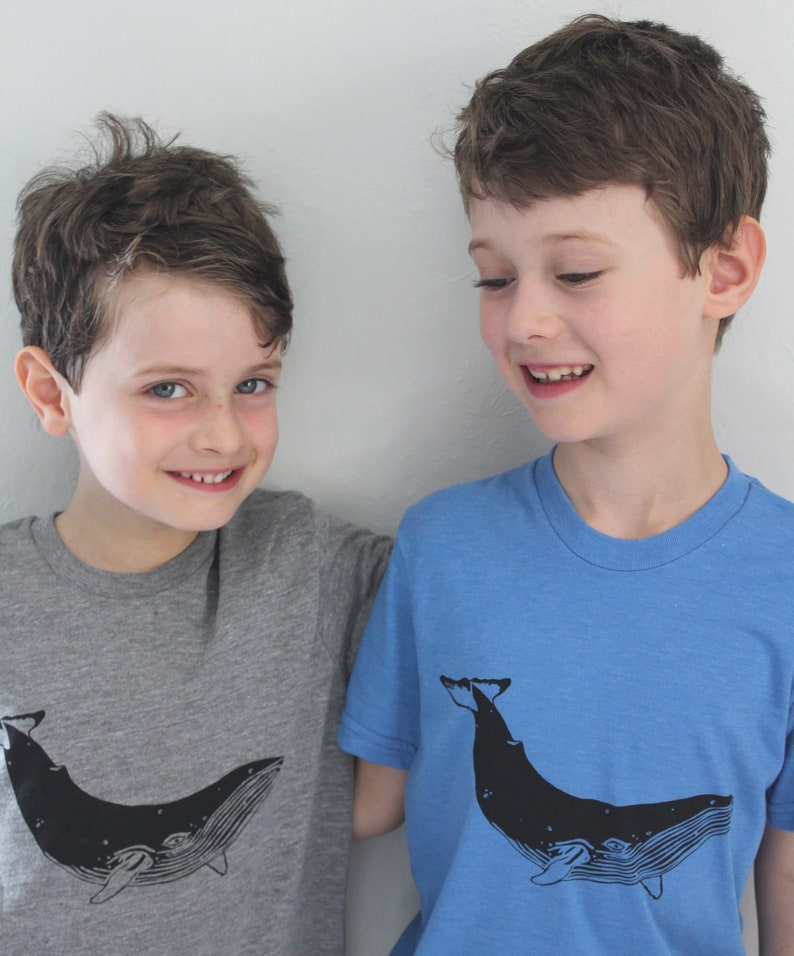 May include: Two boys wearing t-shirts with a whale graphic. The boy on the left is wearing a gray t-shirt with a black whale graphic. The boy on the right is wearing a blue t-shirt with a black whale graphic.