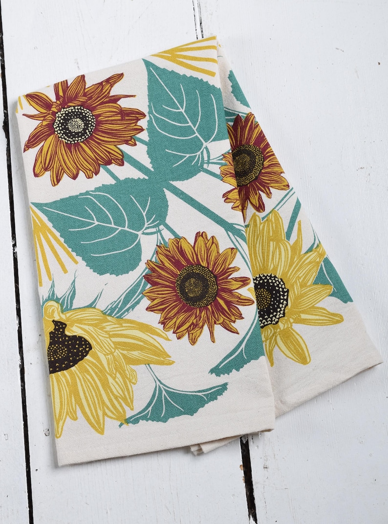 Op de afbeelding: Twee opgevouwen keukendoeken met een zonnebloemontwerp. De handdoeken hebben grote gele en oranje zonnebloemen met donkerbruine centra, tegen een cr&egrave;mekleurige achtergrond met turquoise groene bladeren. De handdoeken zijn gemaakt van een zacht, absorberend materiaal.