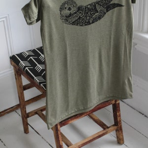 May include: A green t-shirt with a black graphic print of an otter on the back. The shirt is draped over a wooden chair with a black and white patterned seat.