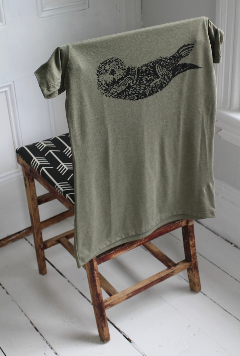 May include: A green t-shirt with a black graphic print of an otter on the back. The shirt is draped over a wooden chair with a black and white patterned seat.