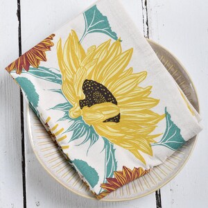 Op de afbeelding: Een gevouwen, cr&egrave;mekleurige keukendoek met een grote gele zonnebloem met een donkerbruin centrum en turquoise groene bladeren. De doek ligt op een beige bord met een geribbeld ontwerp, tegen een witte houten achtergrond.