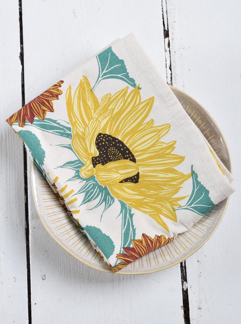 Op de afbeelding: Een gevouwen, cr&egrave;mekleurige keukendoek met een grote gele zonnebloem met een donkerbruin centrum en turquoise groene bladeren. De doek ligt op een beige bord met een geribbeld ontwerp, tegen een witte houten achtergrond.