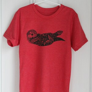 May include: Red t-shirt with a black and white graphic print of a sea otter.