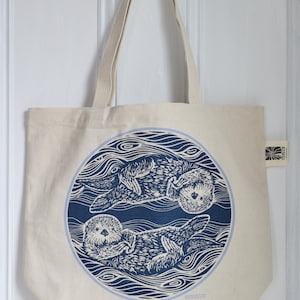 May include: Cream-colored tote bag featuring a blue circular design with two otters swimming in stylized water. The bag has a simple, rectangular shape with long, cream-colored handles for carrying over the shoulder.