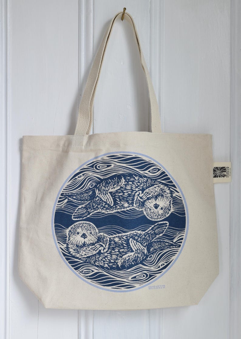 May include: Cream-colored tote bag featuring a blue circular design with two otters swimming in stylized water. The bag has a simple, rectangular shape with long, cream-colored handles for carrying over the shoulder.