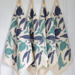 May include: Four off-white kitchen towels hanging from a clothesline, each adorned with a repeating pattern of navy blue and teal sea turtles. The towels are clipped to the line with wooden clothespins, creating a coastal theme.