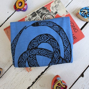 May include: A blue t-shirt with a black and white patterned snake design. The shirt is folded and lying on a white wooden surface.