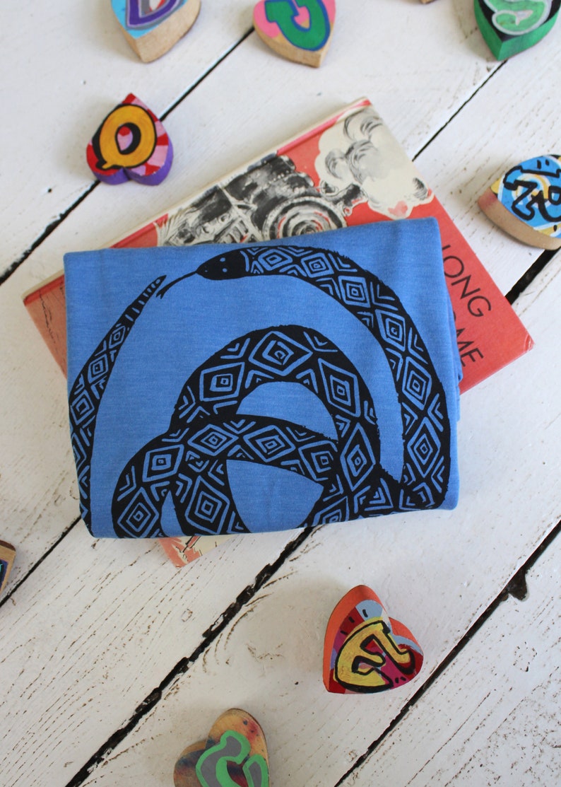 May include: A blue t-shirt with a black and white patterned snake design. The shirt is folded and lying on a white wooden surface.