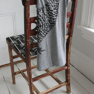 May include: A gray t-shirt with a black and white graphic of a bird printed on it is draped over the back of a wooden chair. The chair has a black and white patterned seat.