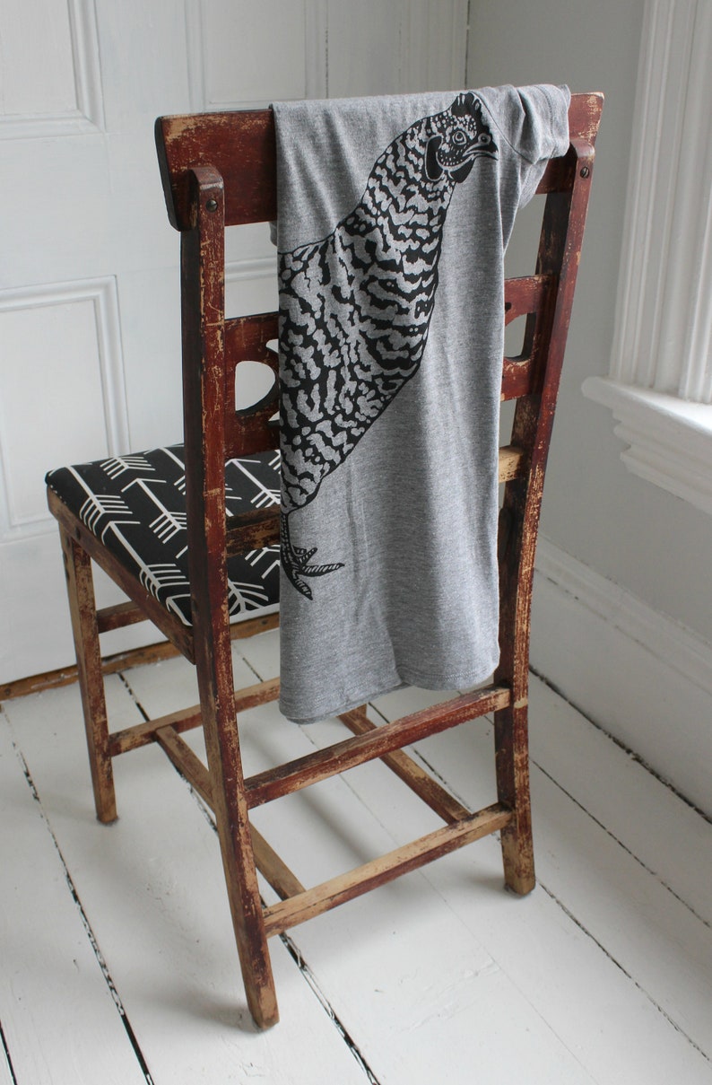 May include: A gray t-shirt with a black and white graphic of a bird printed on it is draped over the back of a wooden chair. The chair has a black and white patterned seat.