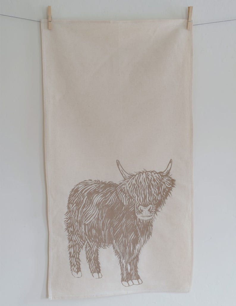 May include: A beige tea towel with a brown illustration of a Highland cow. The towel is hanging from a clothesline with a wooden clothespin. The cow is facing forward with its horns visible.