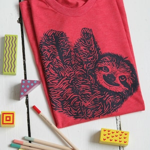 May include: Red t-shirt with a black and white illustration of a sloth. The shirt is made of a soft, comfortable material. The image also shows colored pencils and wooden blocks.