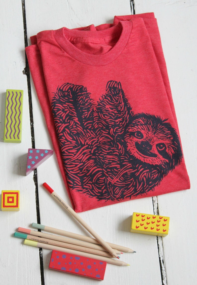 May include: Red t-shirt with a black and white illustration of a sloth. The shirt is made of a soft, comfortable material. The image also shows colored pencils and wooden blocks.