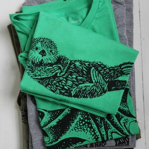 K&ouml;nnte beinhalten: Ein Stapel gefalteter T-Shirts in Gr&uuml;n-, Grau- und Olivt&ouml;nen. Das oberste Shirt ist leuchtend gr&uuml;n mit einer schwarzen Grafik eines Seeotters. Der Markenname "harrow" ist auf dem Kragen gestickt. Die Shirts sind auf einer wei&szlig;en Holzoberfl&auml;che gestapelt.