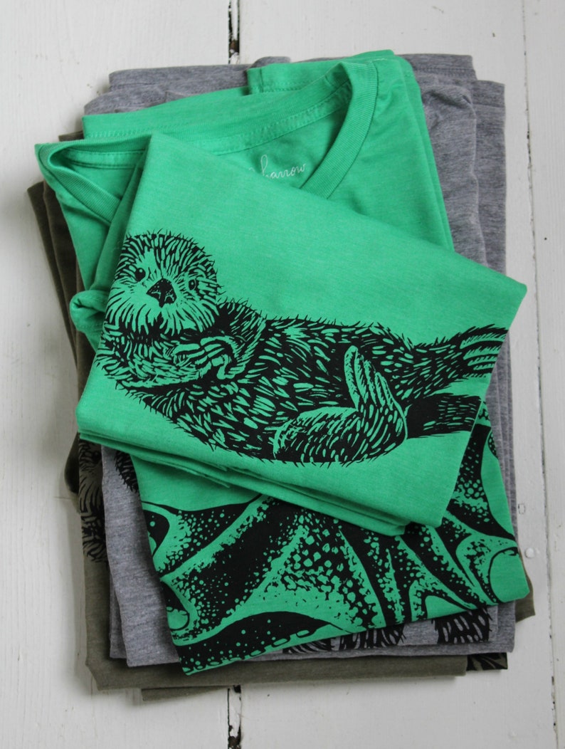 K&ouml;nnte beinhalten: Ein Stapel gefalteter T-Shirts in Gr&uuml;n-, Grau- und Olivt&ouml;nen. Das oberste Shirt ist leuchtend gr&uuml;n mit einer schwarzen Grafik eines Seeotters. Der Markenname "harrow" ist auf dem Kragen gestickt. Die Shirts sind auf einer wei&szlig;en Holzoberfl&auml;che gestapelt.
