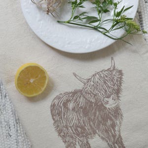 May include: A white linen tea towel with a brown line drawing of a Highland cow. The cow is standing on a white background. Two lemons and a sprig of greenery are on a white plate in the background.