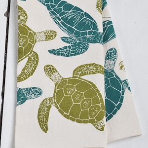 May include: A white kitchen towel with a green and teal sea turtle pattern. The turtles are printed in a block print style.