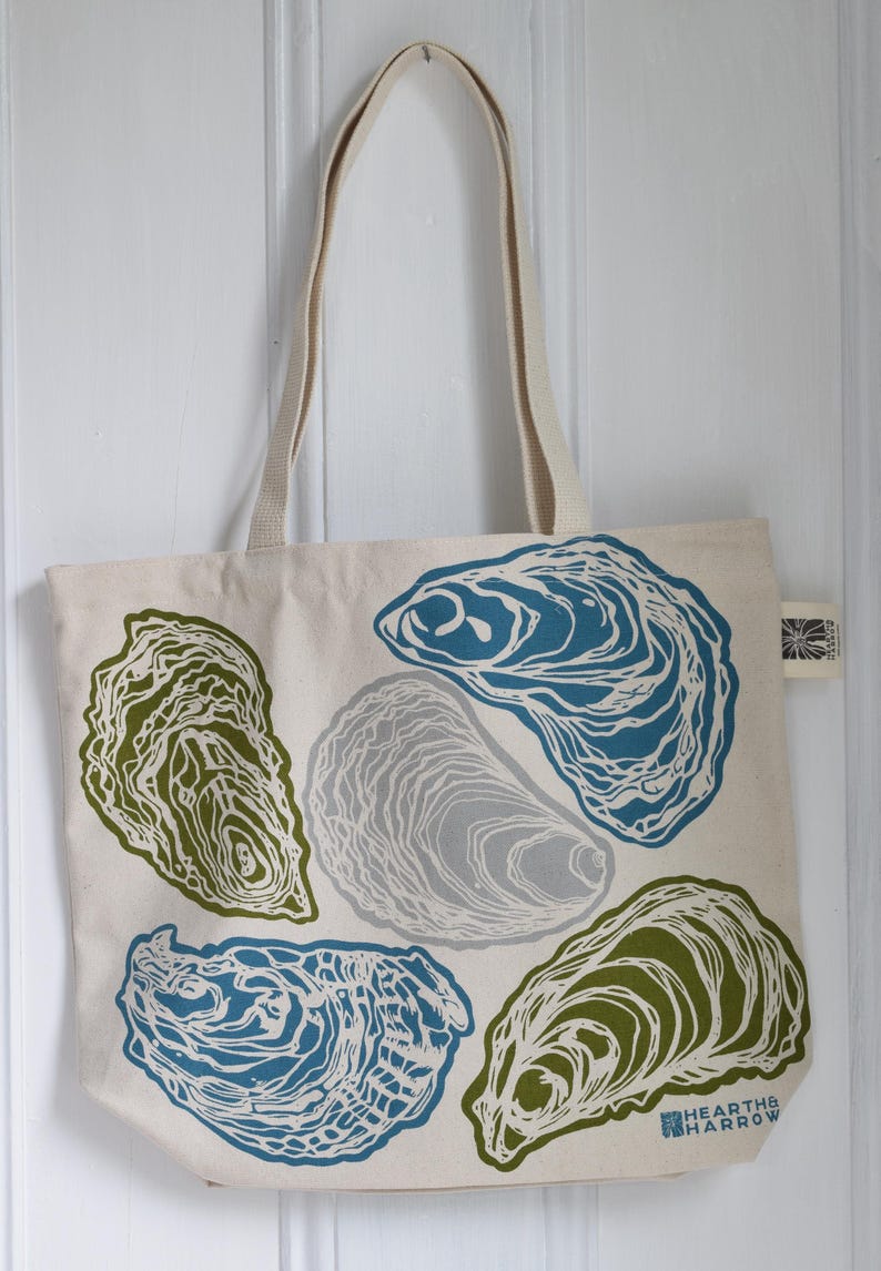 May include: Cream-colored tote bag with a repeating oyster shell design in shades of blue, green, and gray. The bag has long, beige handles and the text "HEARTHE HARROW" is printed on the bottom right corner.