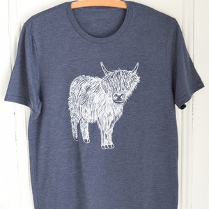 May include: A dark blue t-shirt with a white graphic of a Highland cow. The cow is standing on all four legs and has a long, shaggy coat.