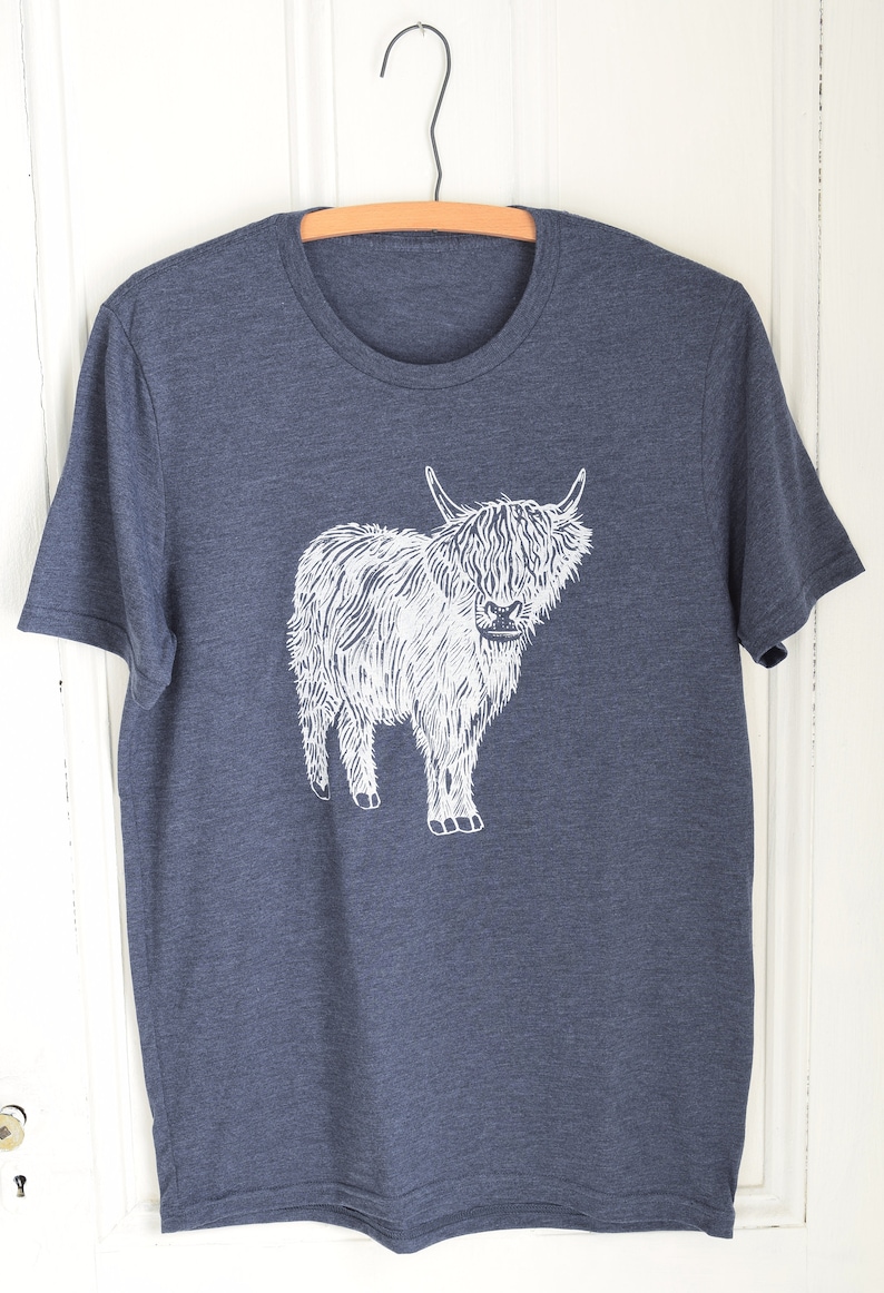 May include: A dark blue t-shirt with a white graphic of a Highland cow. The cow is standing on all four legs and has a long, shaggy coat.