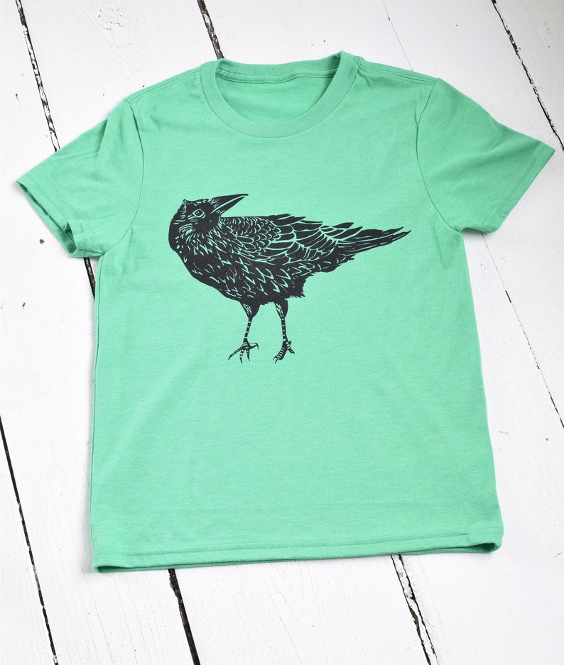 Pode incluir: Camiseta verde claro com uma ilustra&ccedil;&atilde;o preta de um corvo. O corvo &eacute; retratado em um estilo art&iacute;stico detalhado, com suas penas e caracter&iacute;sticas claramente definidas. A camiseta &eacute; feita de um material macio e tem um decote redondo cl&aacute;ssico.