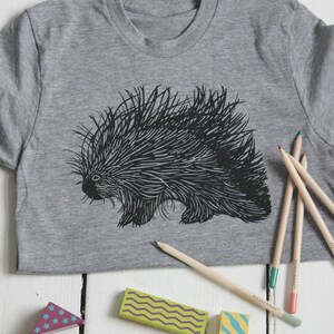 May include: A gray t-shirt with a black and white illustration of a porcupine. The shirt is surrounded by colorful wooden blocks and pencils.