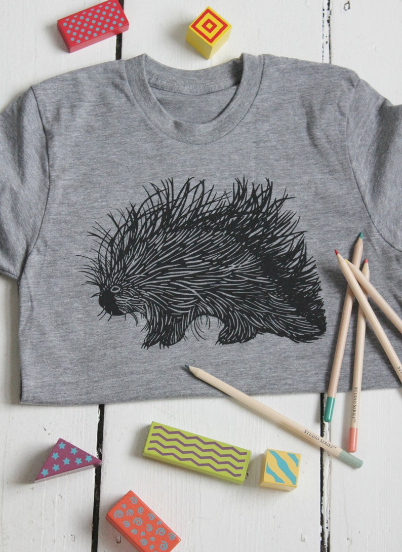 May include: A gray t-shirt with a black and white illustration of a porcupine. The shirt is surrounded by colorful wooden blocks and pencils.