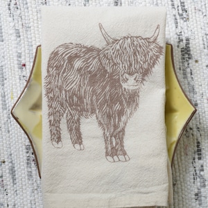 May include: A white kitchen towel with a brown line drawing of a Highland cow. The cow is standing on a white background.