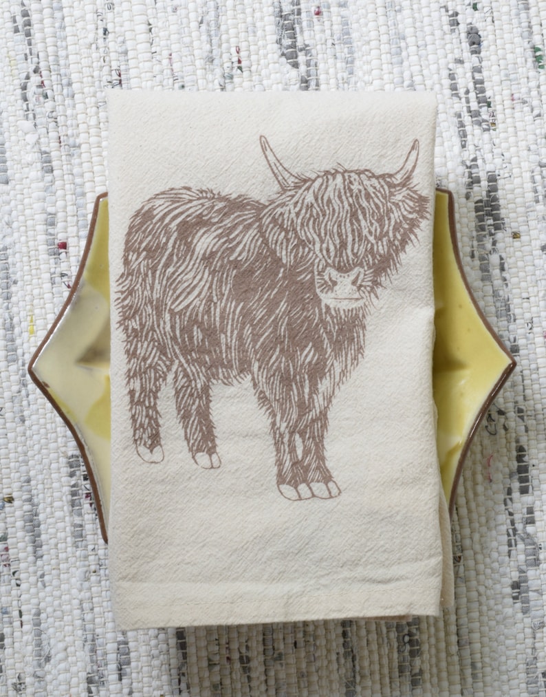 May include: A white kitchen towel with a brown line drawing of a Highland cow. The cow is standing on a white background.